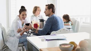 Couple sitting at kitchen table with their 2 young children. Father is showing mother something on his phone. 